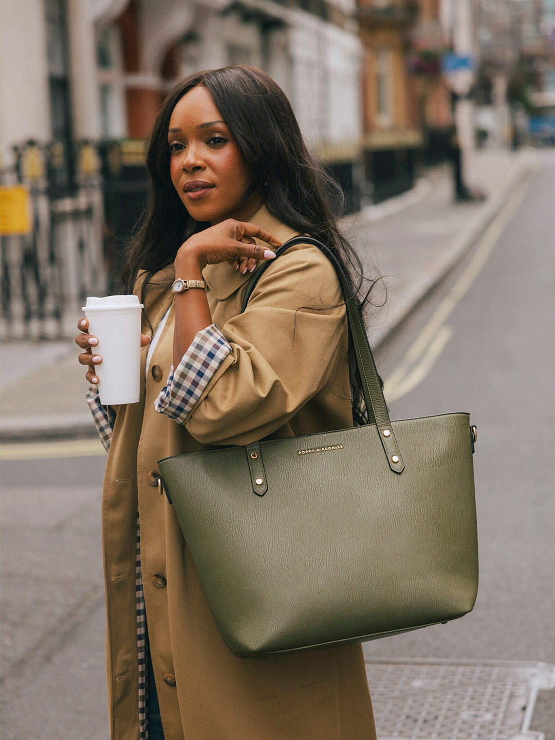 Woman wearing the Poppy & Peonies Go-Getter Tote in Olive over her shoulder while holding a takeaway coffee on a London street.