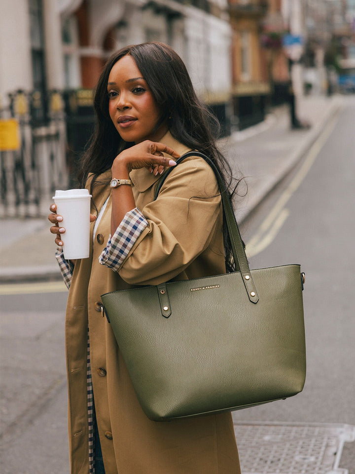 Woman wearing the Poppy & Peonies Go-Getter Tote in Olive over her shoulder while holding a takeaway coffee on a London street.