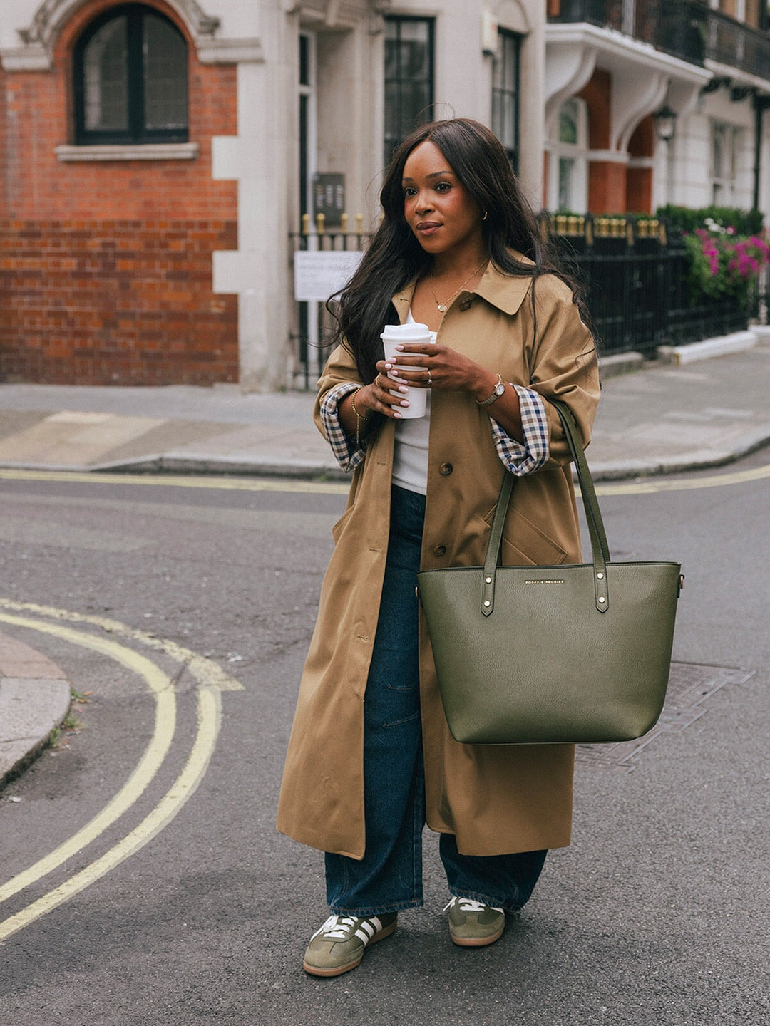 Woman standing with the Poppy & Peonies Go-Getter Tote in Olive carried by the top handles on a London street.