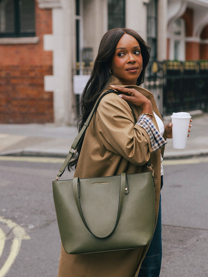 Woman carrying the Poppy & Peonies Go-Getter Tote in Olive over her shoulder while holding a takeaway coffee on a London street.