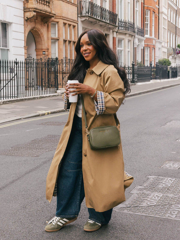 Full-body lifestyle shot of a woman wearing the Poppy & Peonies 24/7 Crossbody in Olive while walking along a city street with a takeaway coffee.