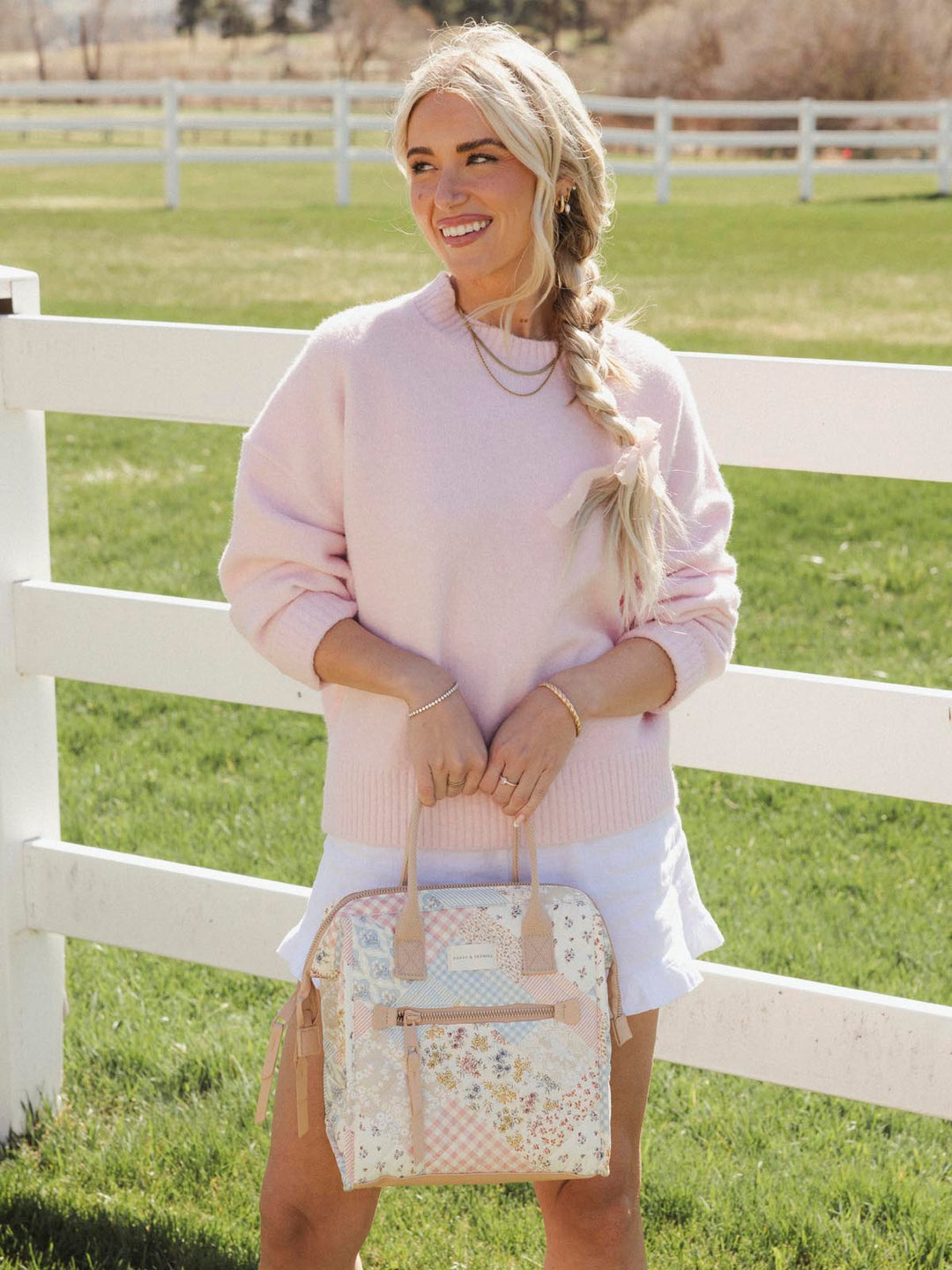 Woman holding a patchwork lunch bag in a grassy field with a white fence.