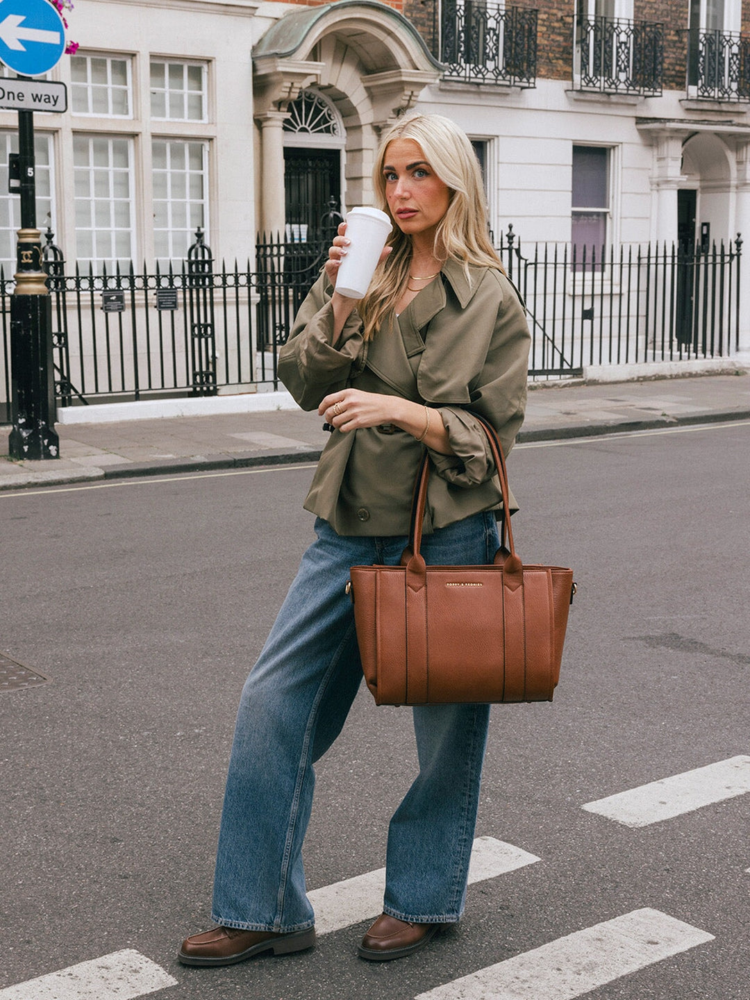 Woman carrying the Poppy & Peonies Small Multitasker in Chestnut by the top handles while holding a takeaway cup.