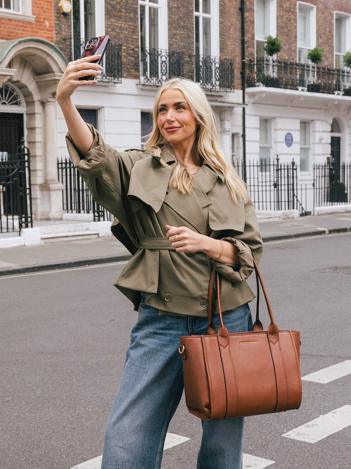 Woman carrying the Poppy & Peonies Small Multitasker in Chestnut by the top handles while taking a selfie on a city street.