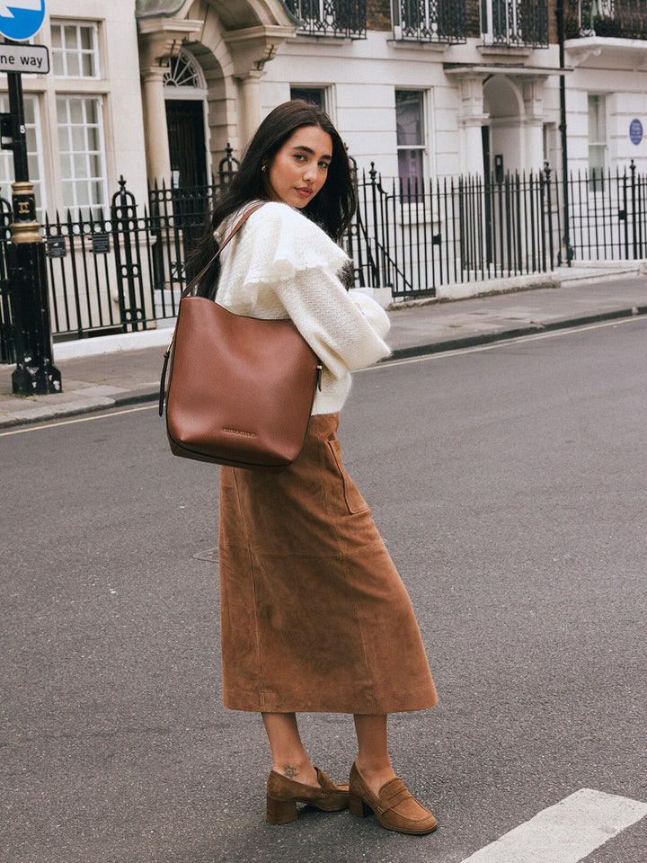 Woman wearing the Poppy & Peonies The Bucket Bag in Chestnut over her shoulder while posing on a city street.
