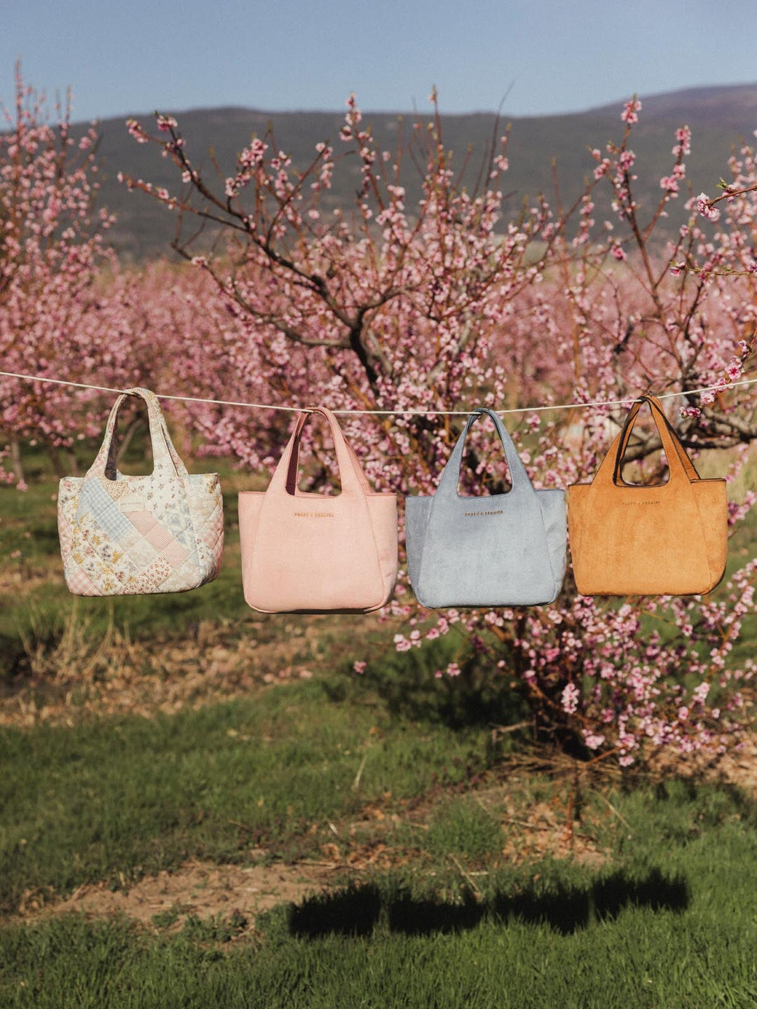 Poppy & Peonies The Pick Me Up Tote in Cognac Suede displayed outdoors with other tote colorways against blooming cherry blossom trees.