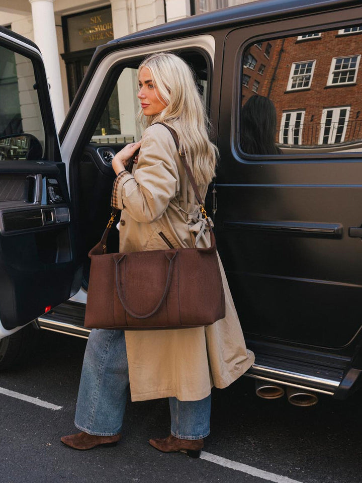 Poppy & Peonies Take Off Tote in Espresso Suede carried by the top handles beside a vehicle, showcasing its roomy travel-ready silhouette.