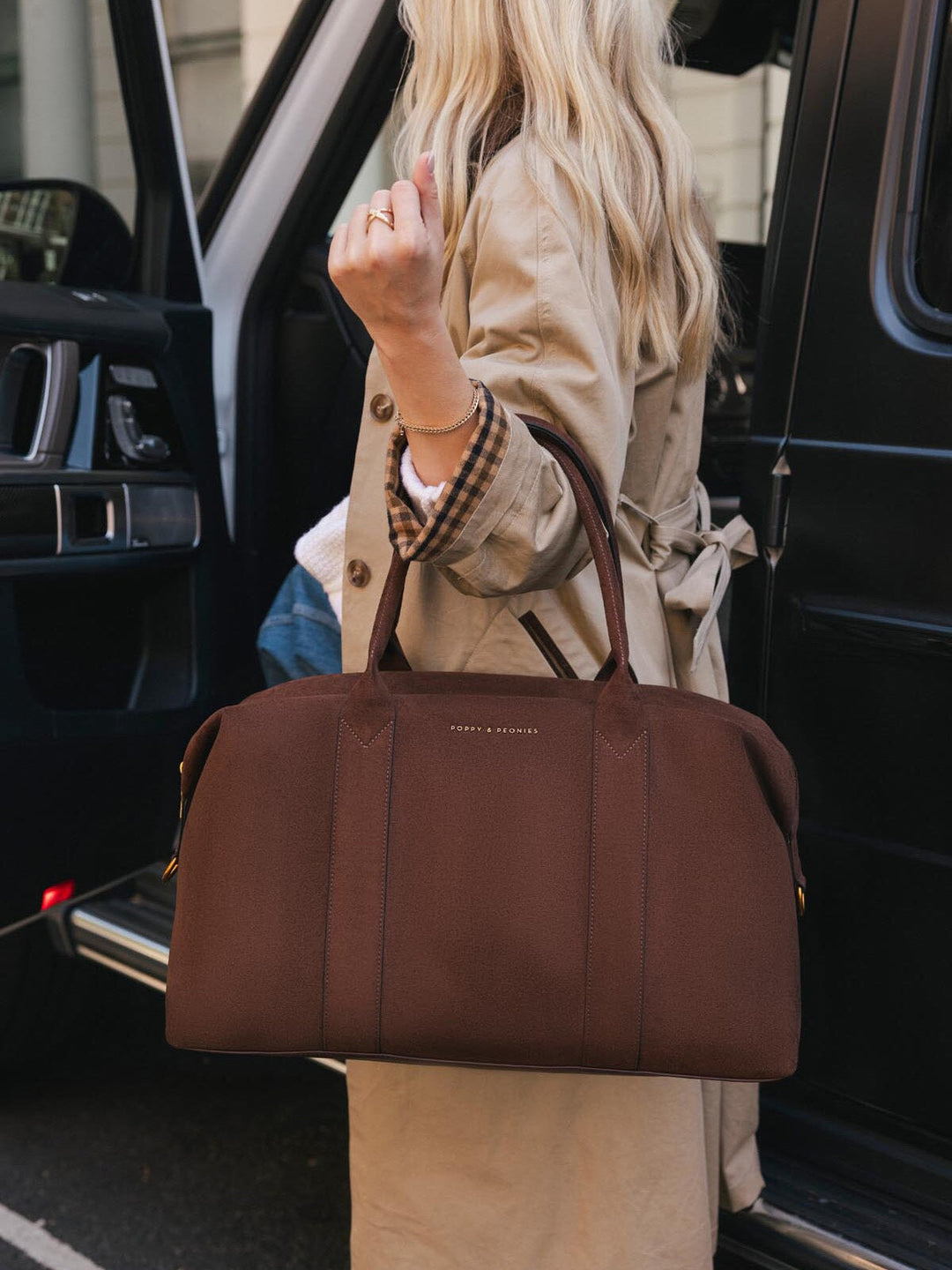 Close-up of Poppy & Peonies Take Off Tote in Espresso Suede carried by hand beside a vehicle, highlighting its spacious silhouette, top handles and soft suede exterior.