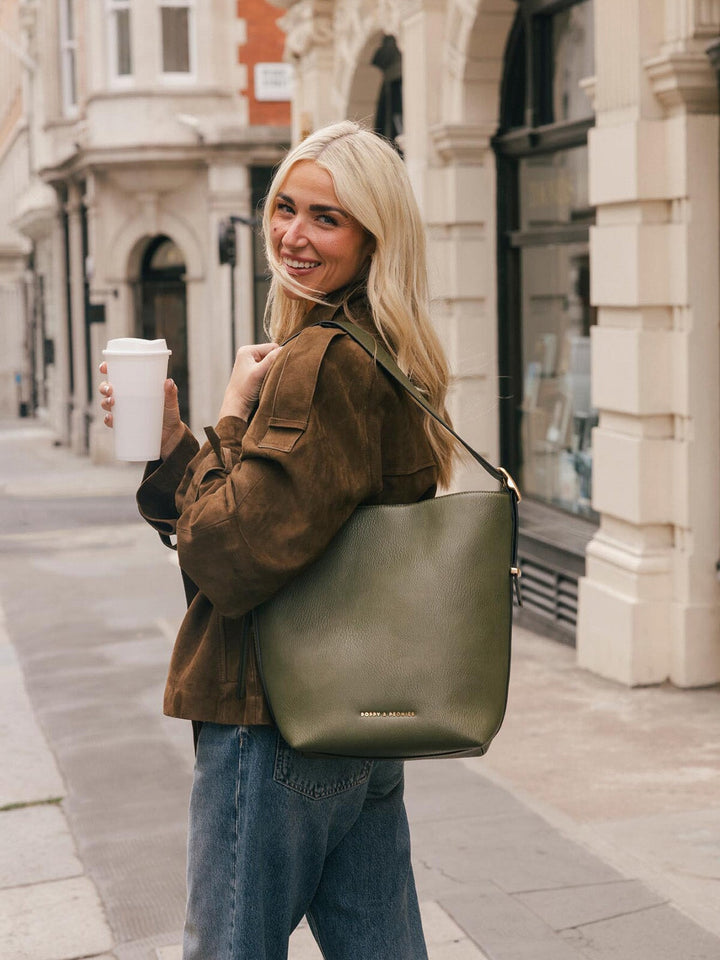 Lifestyle shot of a woman wearing the Poppy & Peonies Bucket Bag in Olive over the shoulder while holding a takeaway coffee.