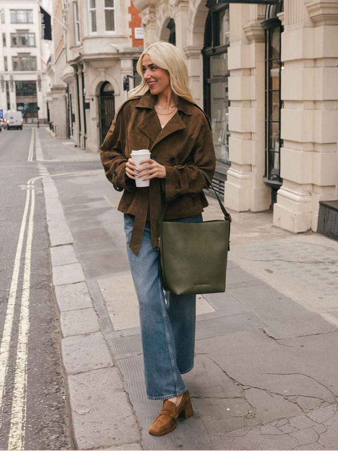 Full-body lifestyle shot of a woman wearing the Poppy & Peonies Bucket Bag in Olive over the shoulder while holding a takeaway coffee.