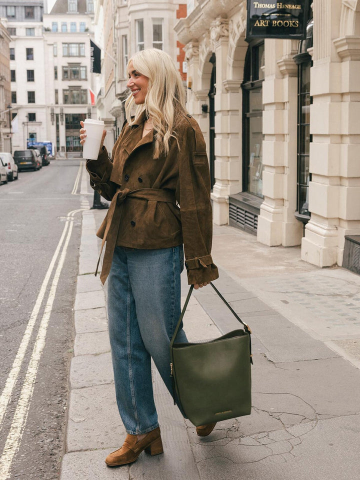 Full-body lifestyle shot of a woman carrying the Poppy & Peonies Bucket Bag in Olive by the shoulder strap while posing on a city street.