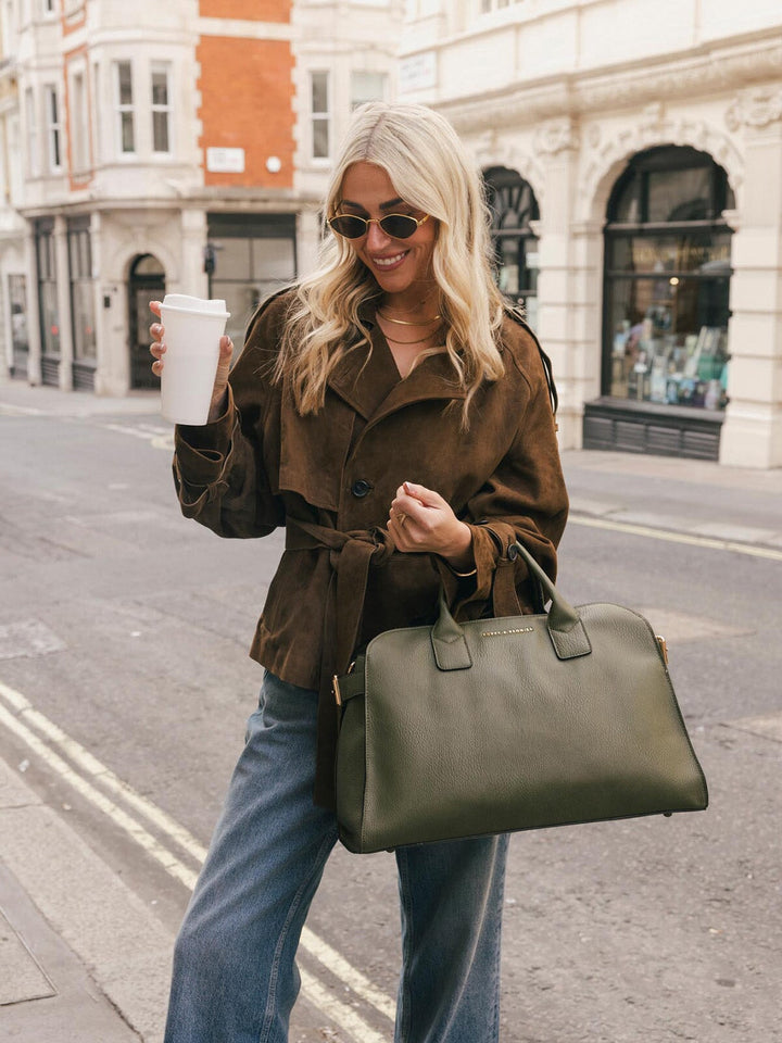 Lifestyle shot of a woman carrying the Poppy & Peonies Uptown Tote in Olive by the top handles while holding a takeaway coffee.