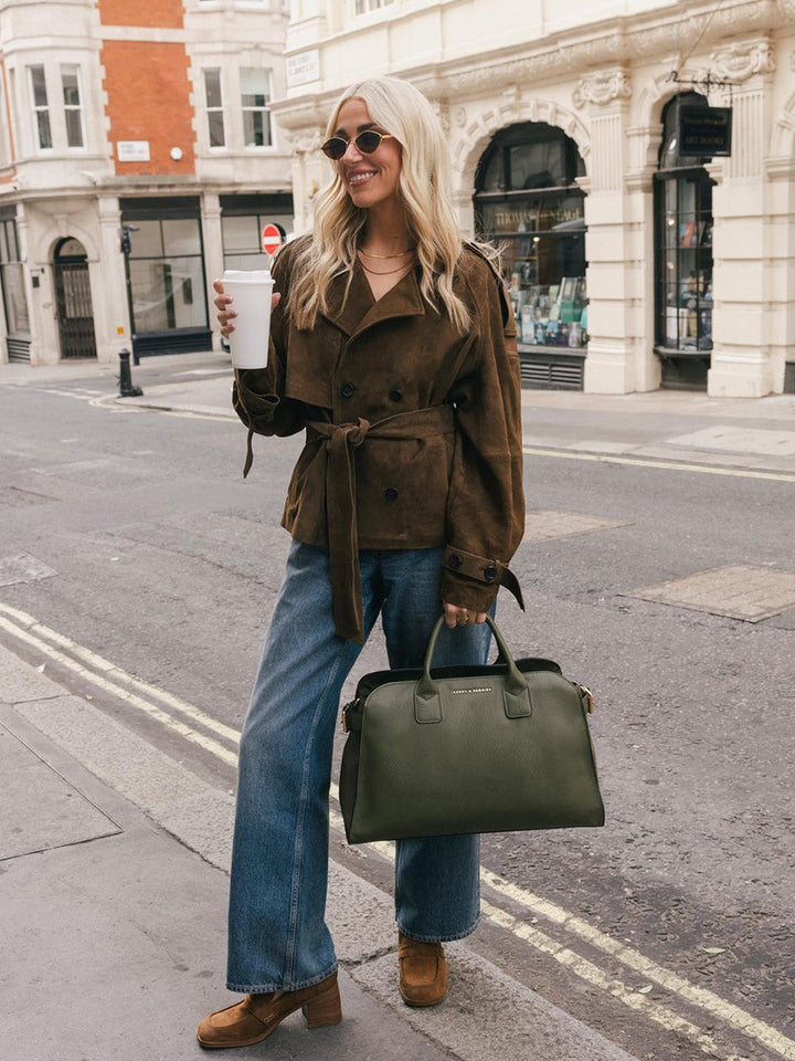 Full-body lifestyle shot of a woman carrying the Poppy & Peonies Uptown Tote in Olive by the top handles while walking along a city street.