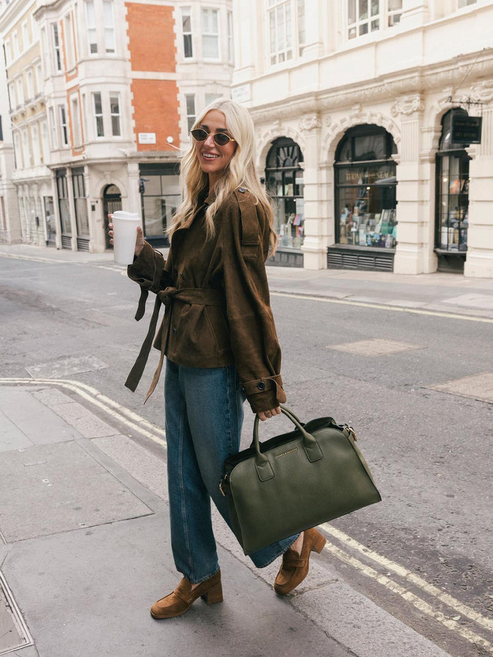 Full-body lifestyle shot of a woman carrying the Poppy & Peonies Uptown Tote in Olive by the top handles while holding a takeaway coffee.