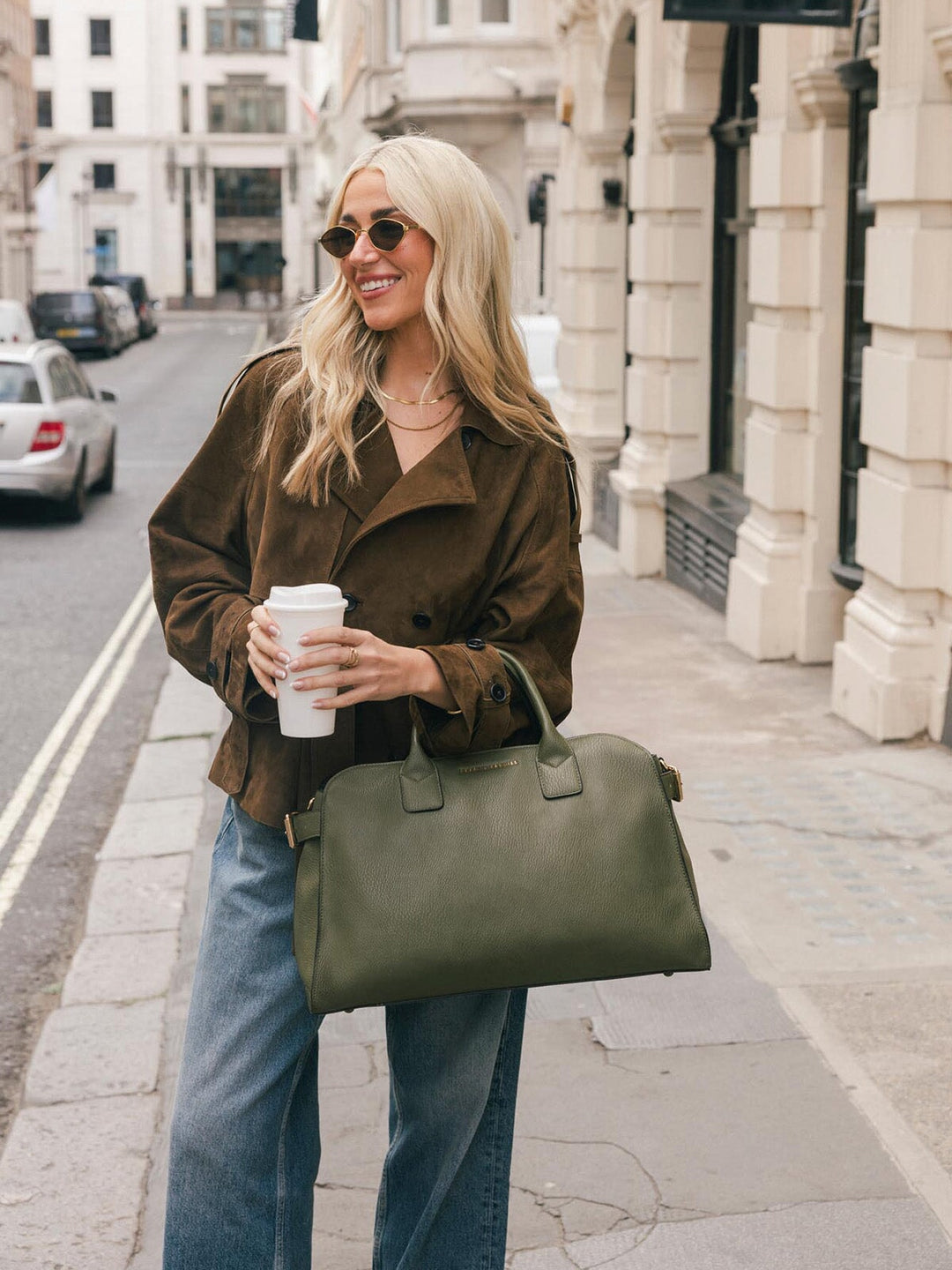 Lifestyle shot of a woman carrying the Poppy & Peonies Uptown Tote in Olive by the top handles while holding a takeaway coffee.