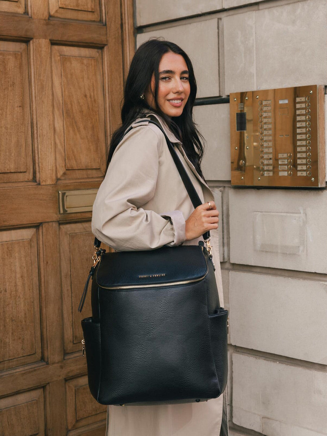 Woman wearing the Poppy & Peonies Carryall Backpack in Black over one shoulder while standing outside a city building.
