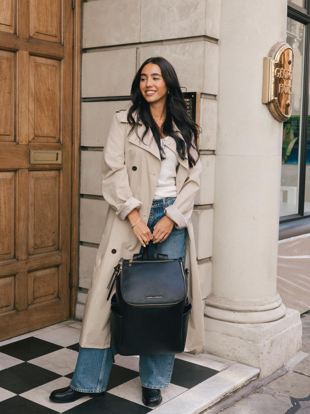 Woman carrying the Poppy & Peonies Carryall Backpack in Black by the top handle while standing outside a city building.
