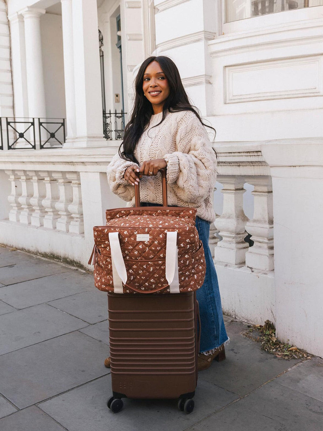 Woman standing with the Poppy & Peonies Mini Voyager in Brown Floral secured over the handle of a rolling suitcase.