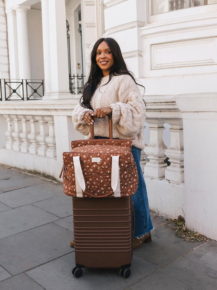 Woman standing with the Poppy & Peonies Mini Voyager in Brown Floral secured over the handle of a rolling suitcase.