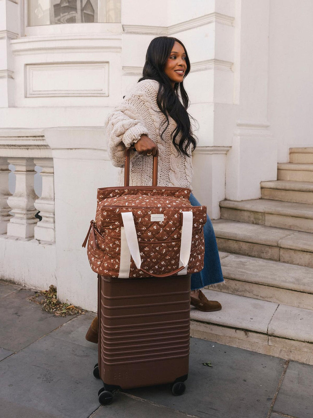 Woman standing beside a rolling suitcase with the Poppy & Peonies Mini Voyager in Brown Floral secured over the luggage handle.