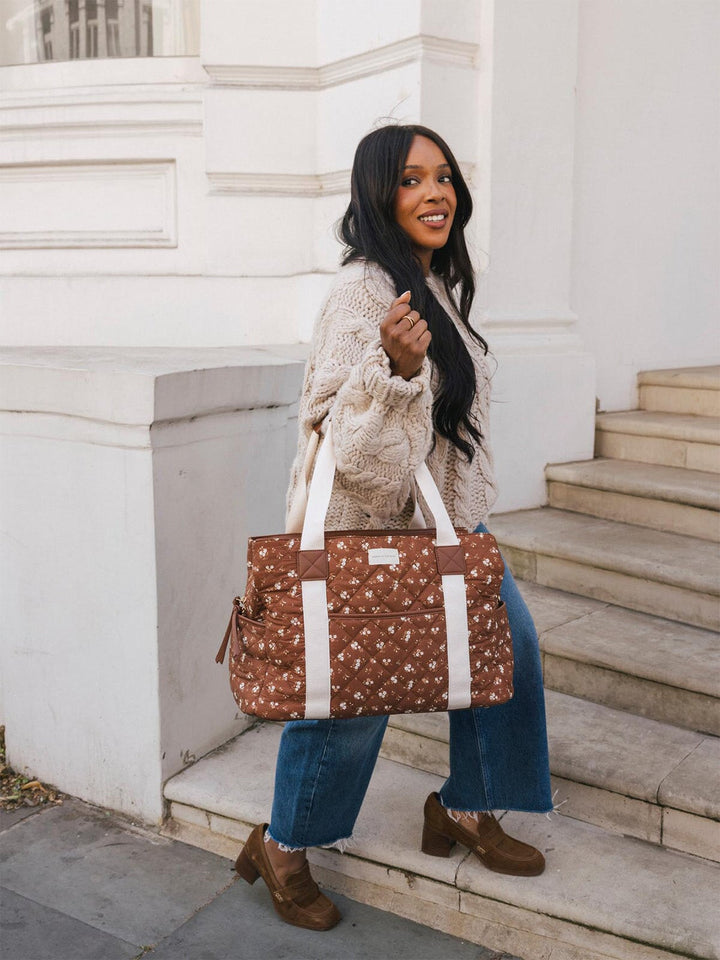 Lifestyle shot of a woman carrying the Poppy & Peonies Mini Voyager in Brown Floral, showcasing its quilted floral patchwork exterior, roomy silhouette, front zip pocket, side pockets, and contrasting shoulder straps.
