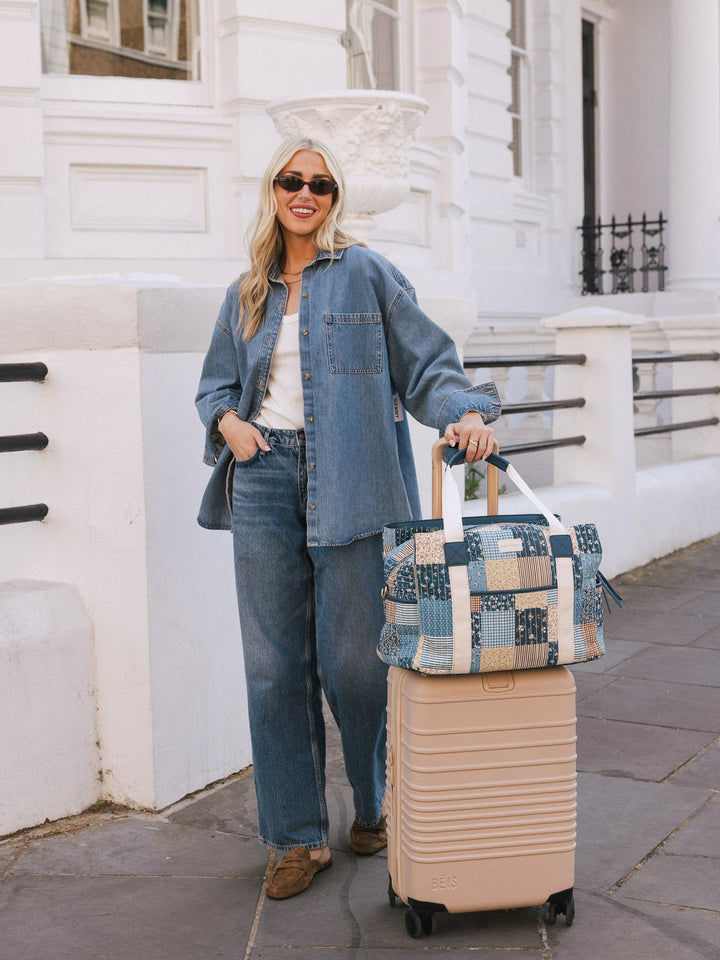 Woman posing with the Poppy & Peonies Mini Voyager in Navy Patchwork secured over the handle of a rolling suitcase.