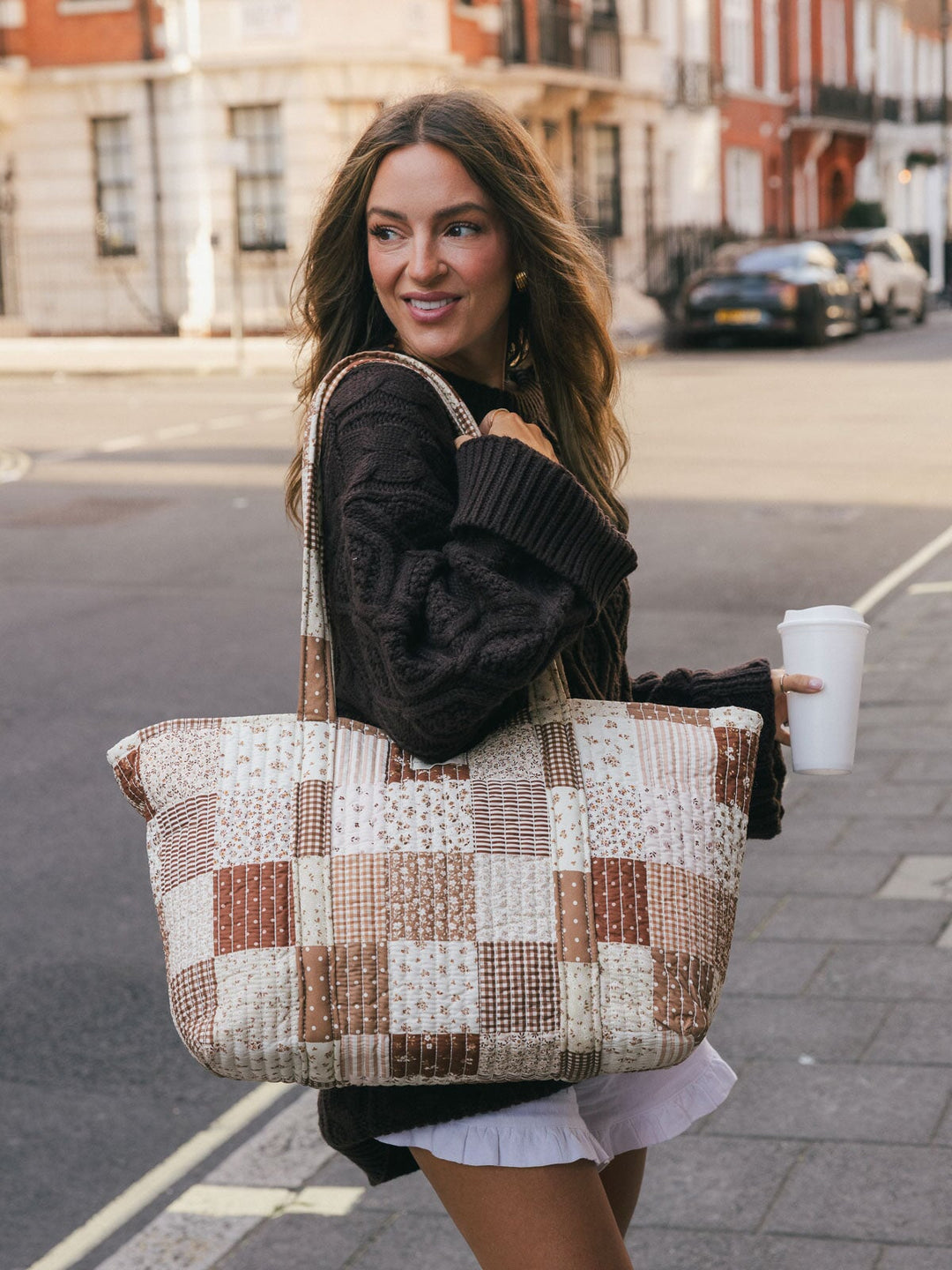 Woman carrying the Poppy & Peonies Daydreamer Tote in Brown Patchwork over her shoulder while holding a takeaway coffee on a city street.