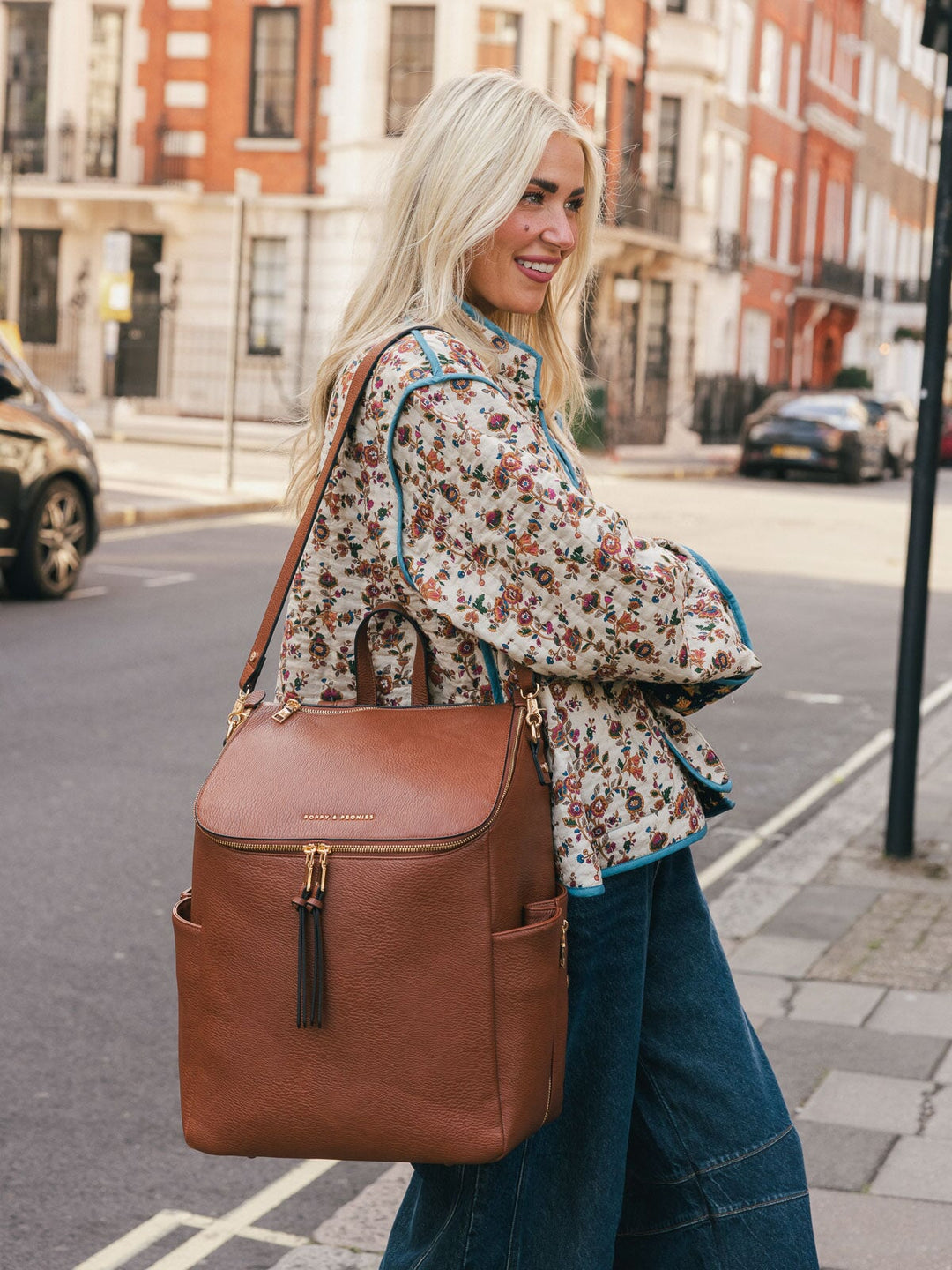 Woman carrying the Poppy & Peonies Carryall Backpack in Chestnut over one shoulder while smiling on a city street.