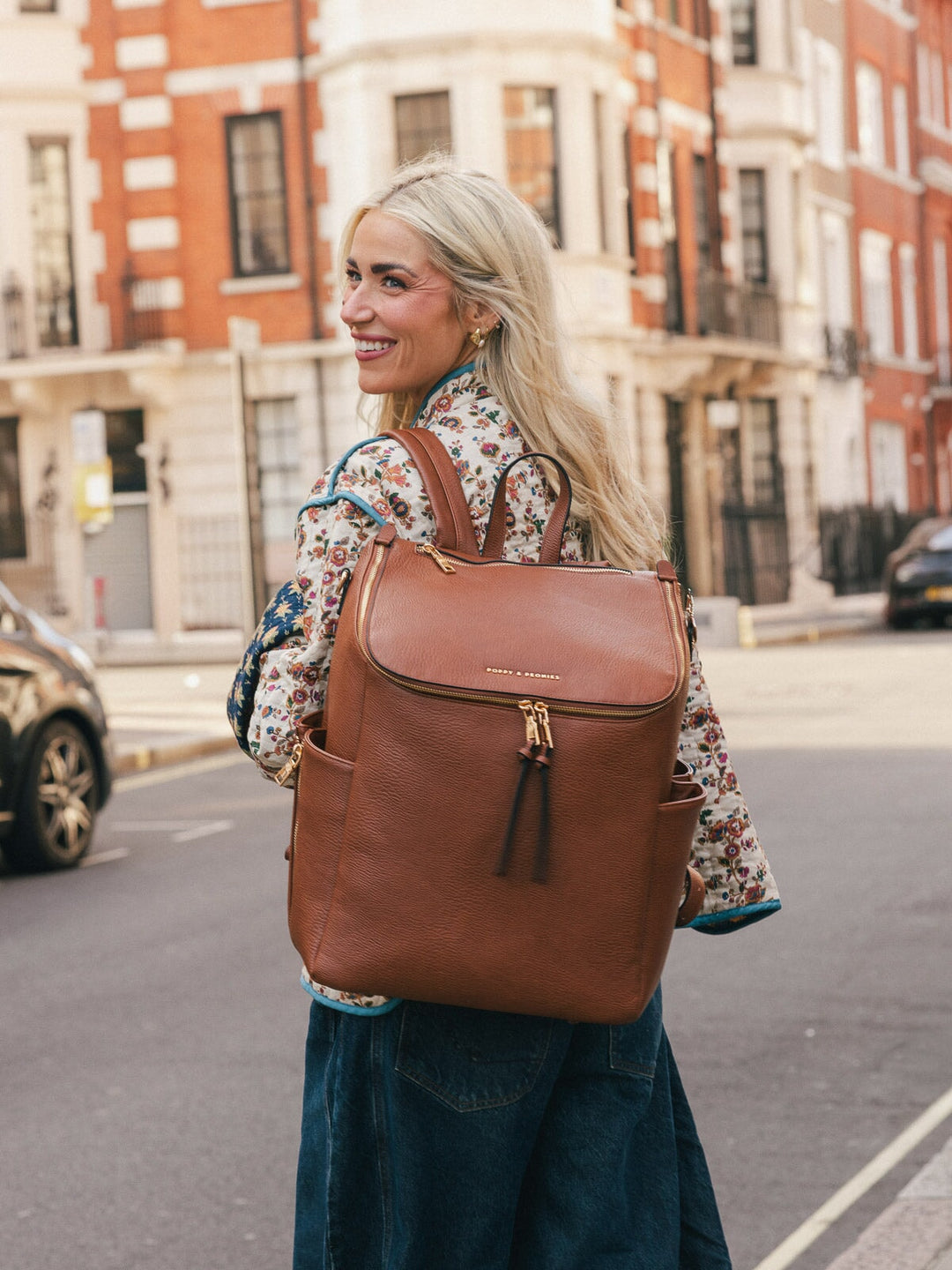 Woman wearing the Poppy & Peonies Carryall Backpack in Chestnut on her back while looking over her shoulder outdoors.