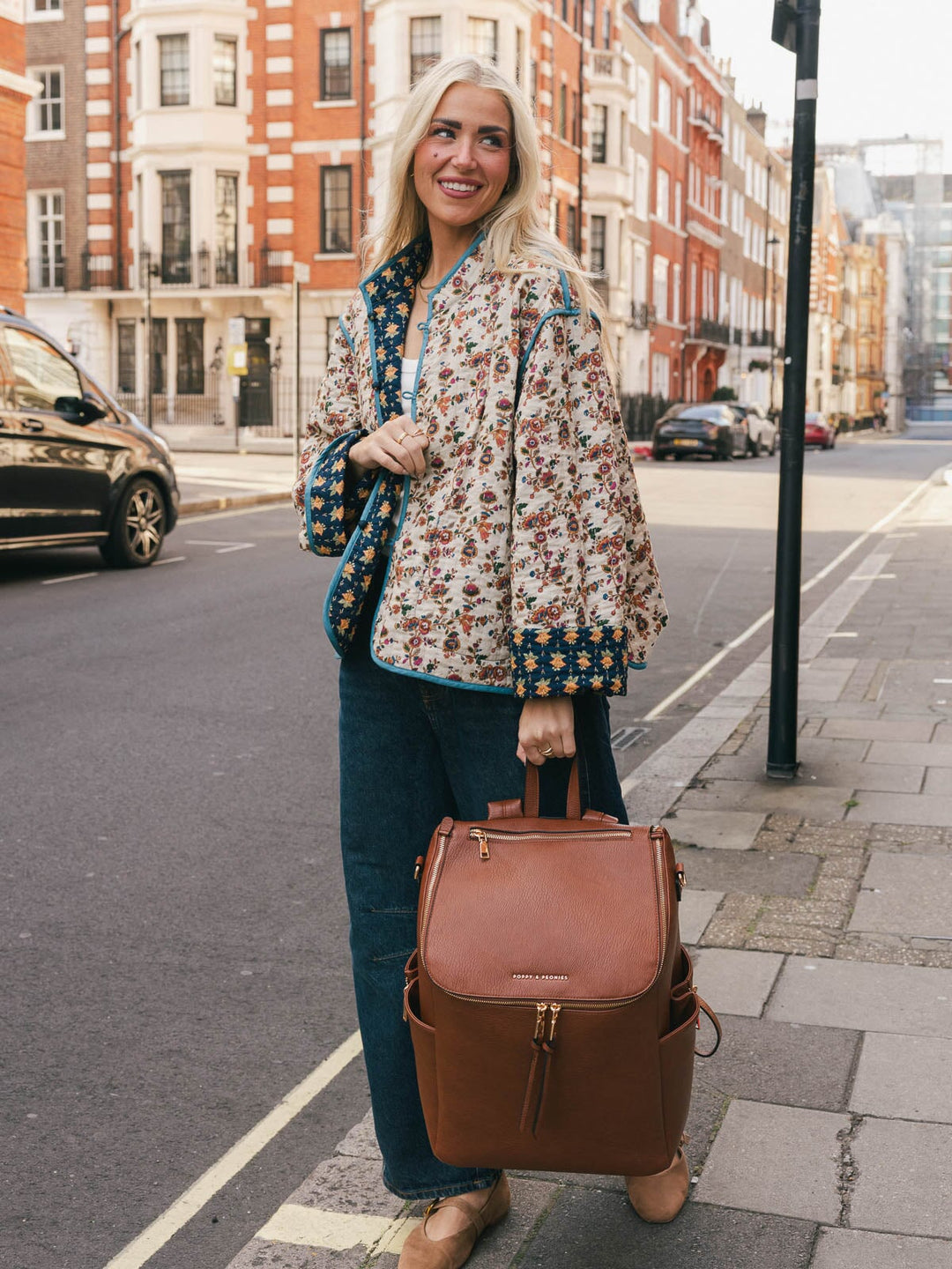 Woman holding the Poppy & Peonies Carryall Backpack in Chestnut by the top handle while smiling outdoors.