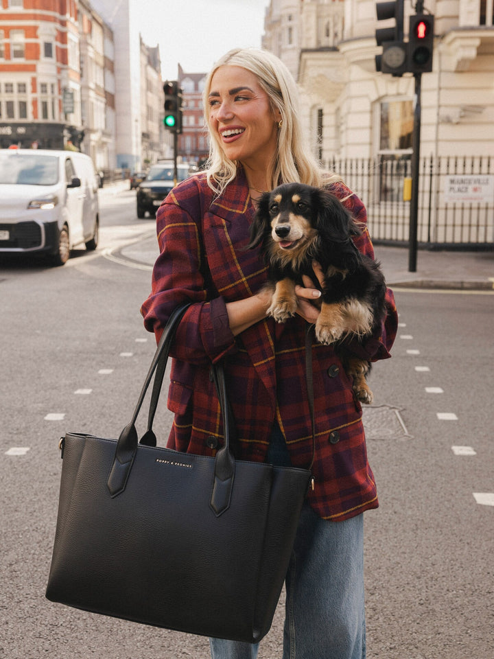 Close-up of a woman carrying the Poppy & Peonies Double Duty Tote in Black by the top handles while holding a small dog.