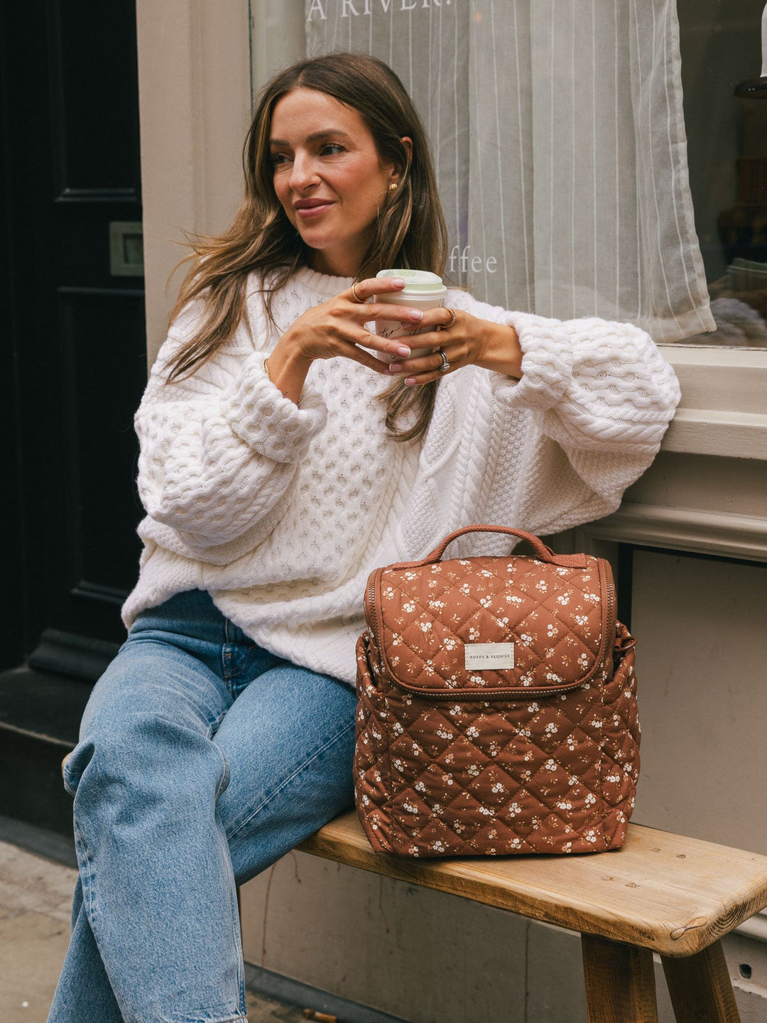 Seated lifestyle shot of a woman holding a takeaway coffee with the Poppy & Peonies Lunch Date in Brown Floral displayed beside her.