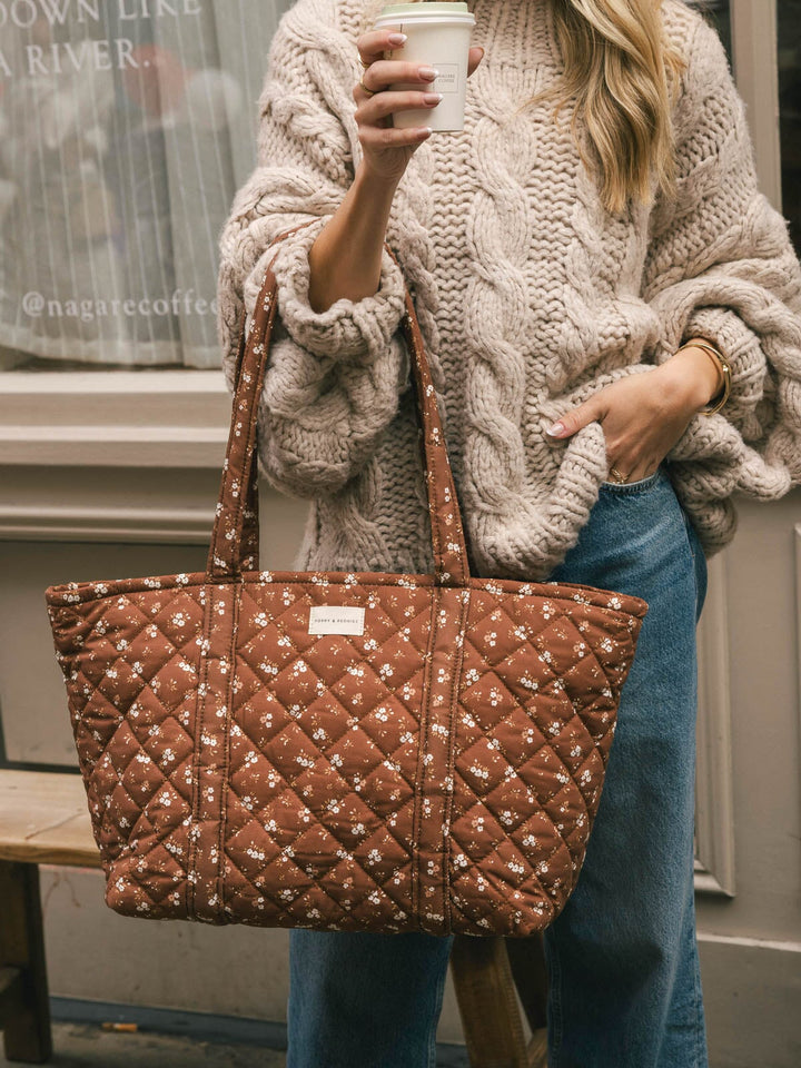 Close-up lifestyle shot of a woman holding a takeaway coffee with the Poppy & Peonies Daydreamer Tote in Brown Floral carried by its handles.
