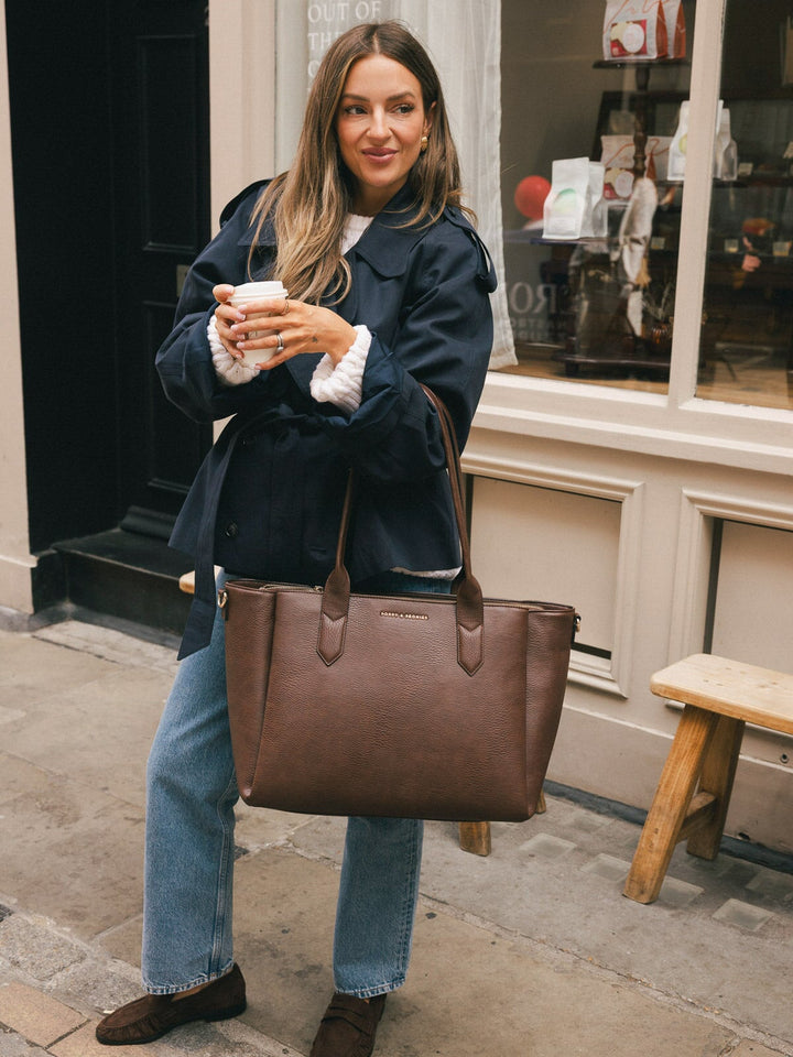 Woman standing with the Poppy & Peonies Double Duty Tote in Espresso held by the top handles while holding a takeaway coffee.