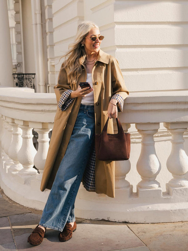 Poppy & Peonies The Pick Me Up Tote in Espresso Suede carried by hand, styled with a trench coat and jeans outdoors.
