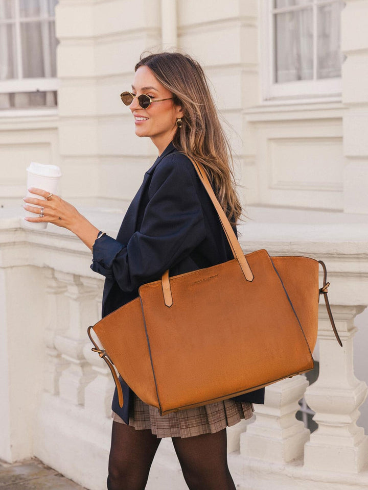Poppy & Peonies Avenue Tote in Cognac Suede carried over the shoulder while holding a coffee, highlighting its roomy everyday silhouette.