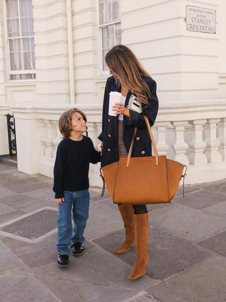 Poppy & Peonies Avenue Tote in Cognac Suede worn over the shoulder during a city walk with a child, highlighting its spacious silhouette and long top handles.