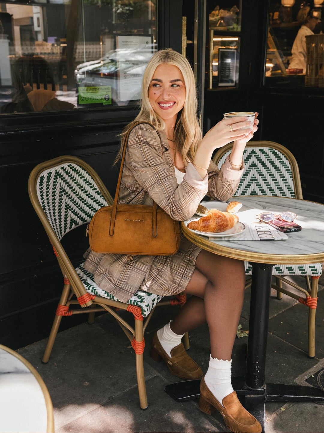 Poppy & Peonies The Friday Satchel in Cognac Suede carried by the top handles while seated at an outdoor café, highlighting its east-west silhouette and warm suede finish.