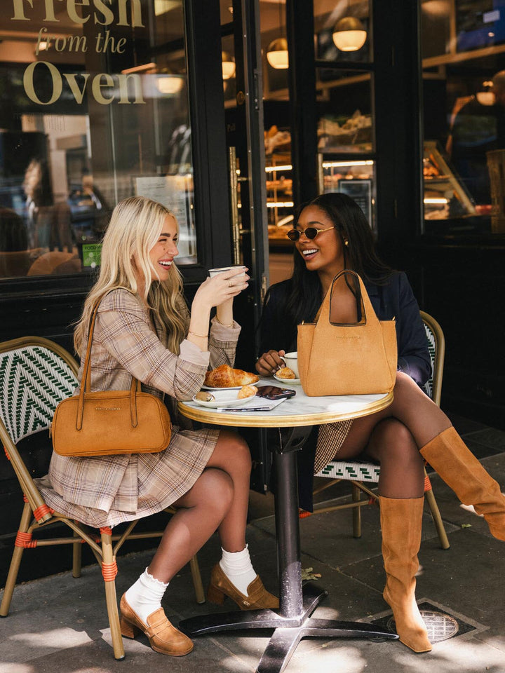 Poppy & Peonies The Pick Me Up Tote in Cognac Suede displayed at an outdoor café, showing its compact structured silhouette and top handles.