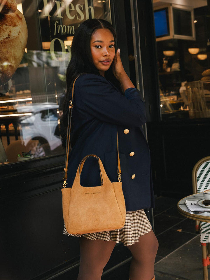 Poppy & Peonies The Pick Me Up Tote in Cognac Suede carried at the side outside a café, showcasing its top handles and everyday size.
