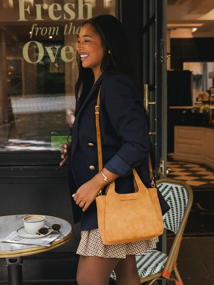 Poppy & Peonies The Pick Me Up Tote in Cognac Suede carried at the side outside a café.