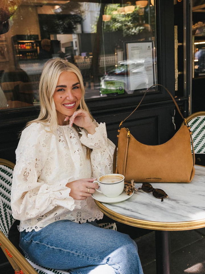 Poppy & Peonies The Sunday Bag in Cognac Suede displayed on an outdoor café table, showing its curved silhouette and shoulder strap.