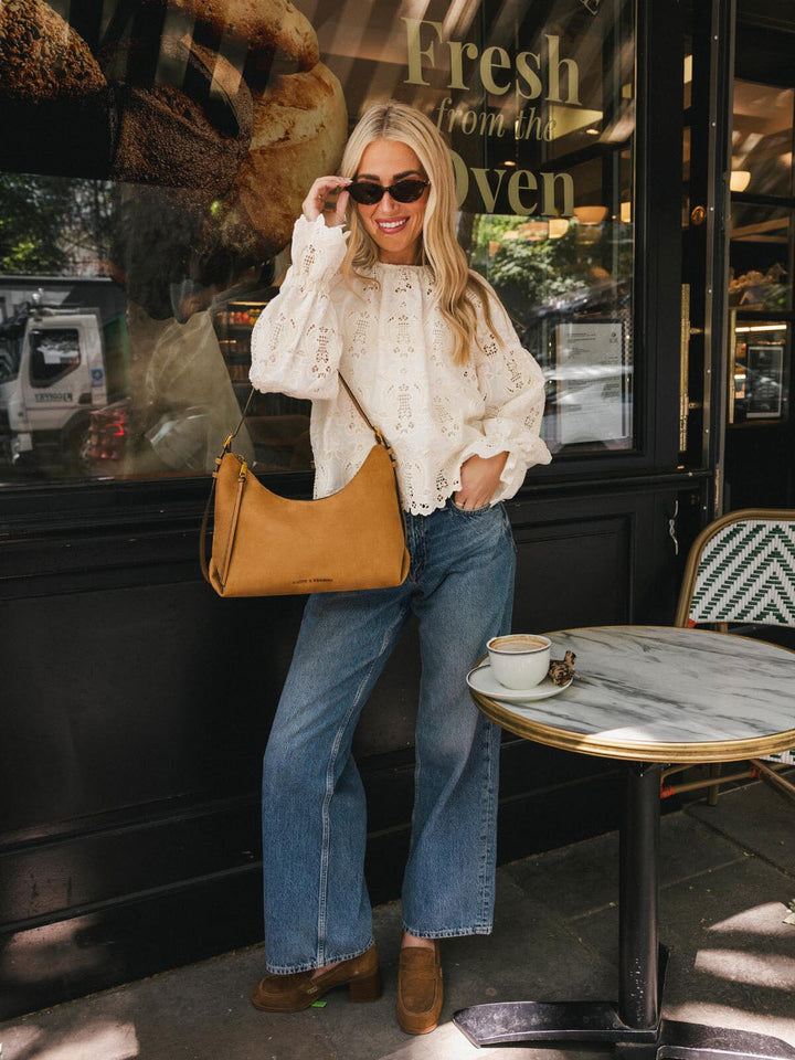 Poppy & Peonies The Sunday Bag in Cognac Suede worn over the shoulder outside a café, styled with a white eyelet blouse and jeans.