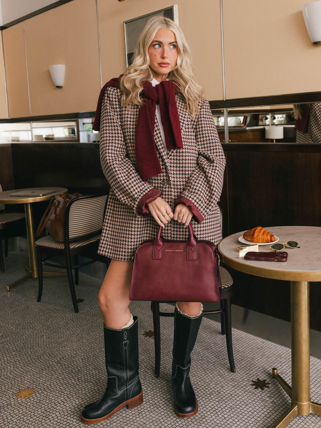 Full-body lifestyle shot of a woman holding the Poppy & Peonies Mini Uptown Tote in Berry by its top handles beside a café table.