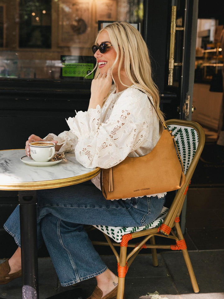 Poppy & Peonies The Sunday Bag in Cognac Suede worn over the shoulder while seated at an outdoor café.