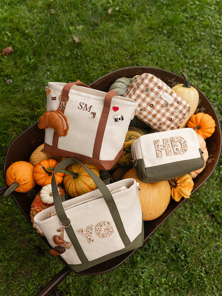 Poppy & Peonies The Small Signature Tote in Olive and Chestnut displayed with coordinating personalized cosmetic case and accessories in a rustic wheelbarrow surrounded by pumpkins and gourds.