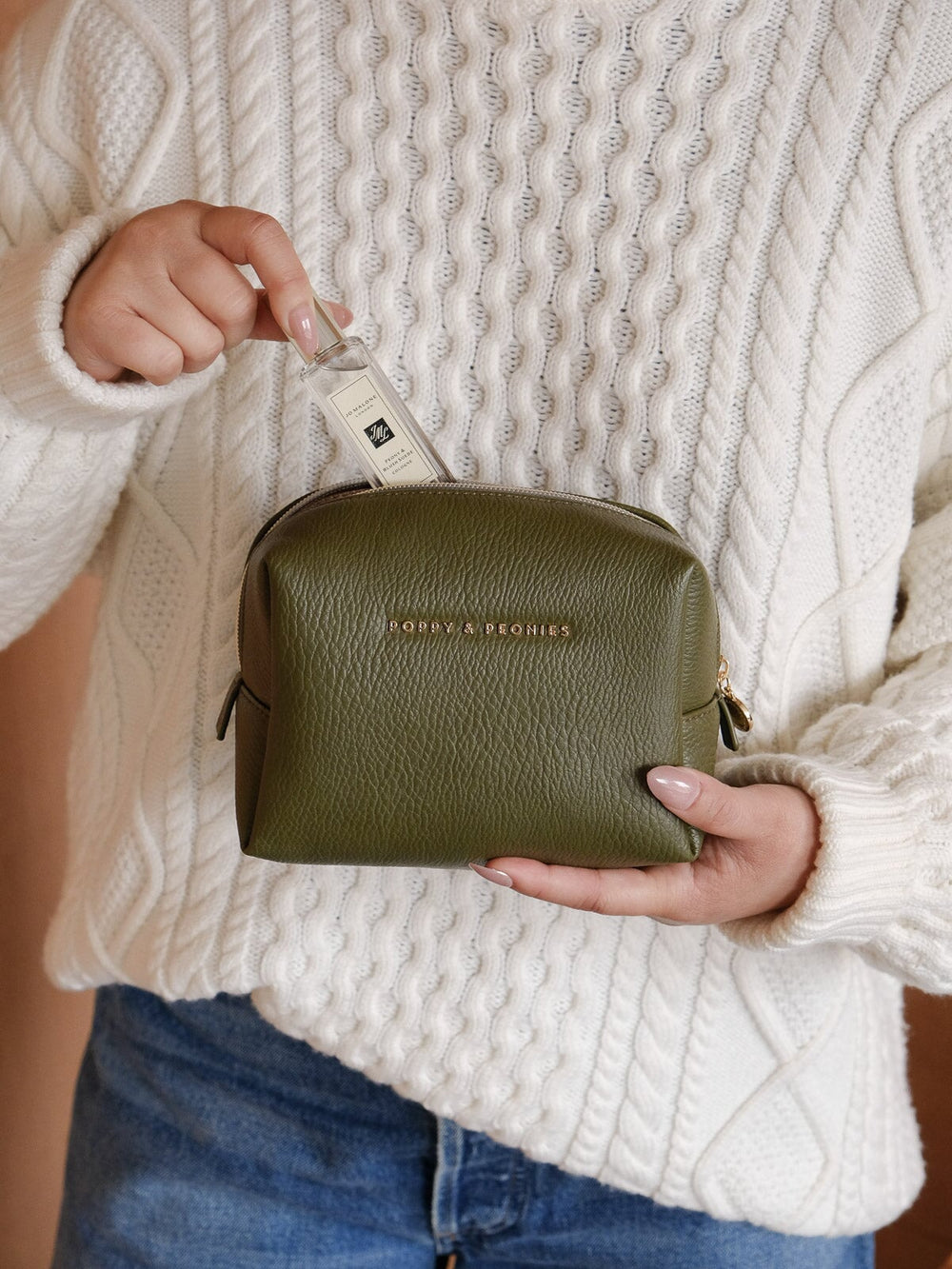 Lifestyle shot of a woman holding the Poppy & Peonies Cosmetic Case in Olive, showcasing its compact shape, textured finish, top handle, and zip closure.