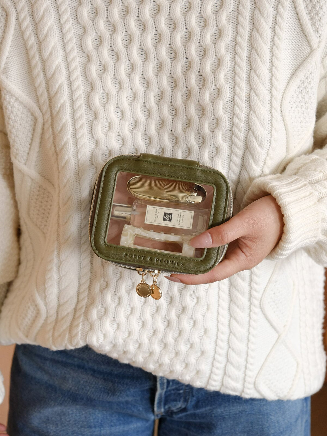 Lifestyle shot of the Mini I See You Cosmetic Case in Olive featuring its clear front panel, compact design, olive trim, and gold-tone zip closures.