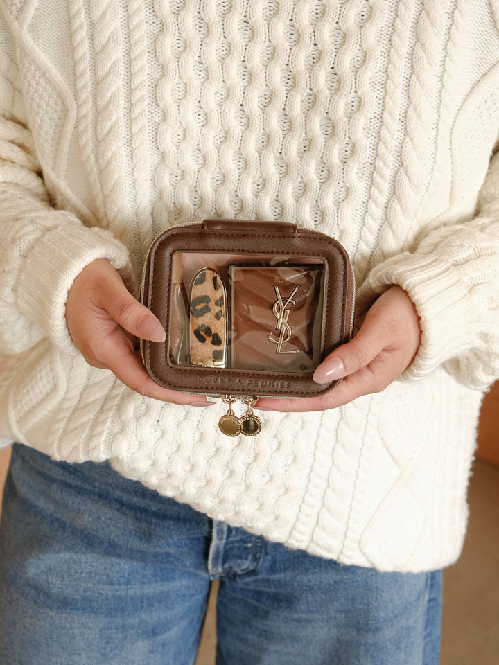 Lifestyle shot of the Mini I See You Cosmetic Case in Espresso featuring its clear front panel with beauty essentials visible inside and gold-tone zip closures.