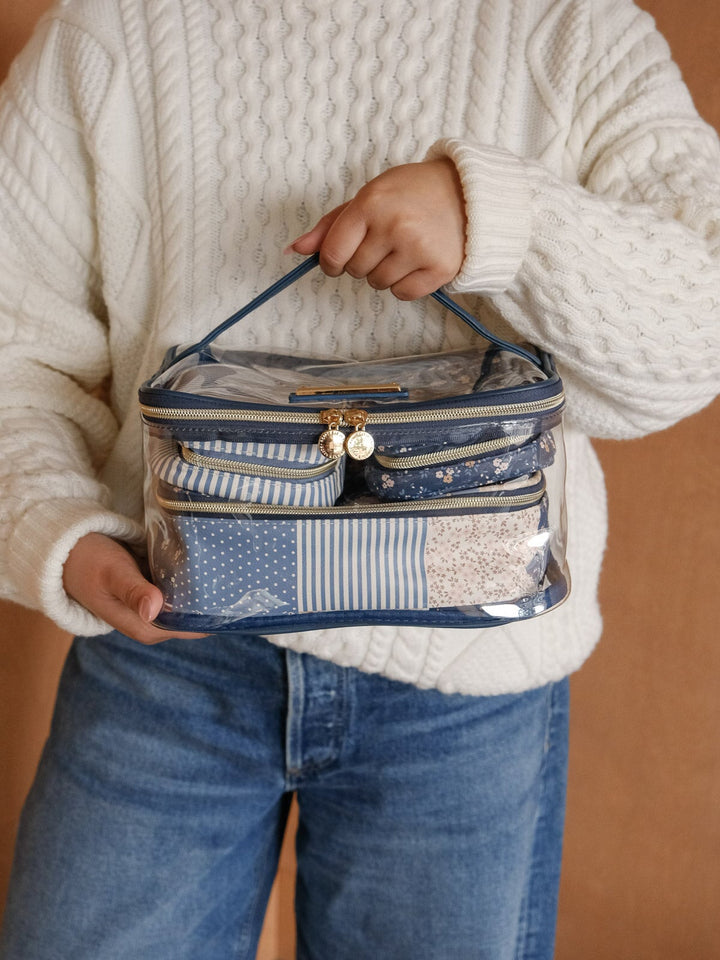 Lifestyle shot of a woman holding the Poppy & Peonies Getaway Cosmetic Set in Navy Patchwork, showcasing its blue patchwork print, clear compartments, top handle, and gold-tone zip closures.